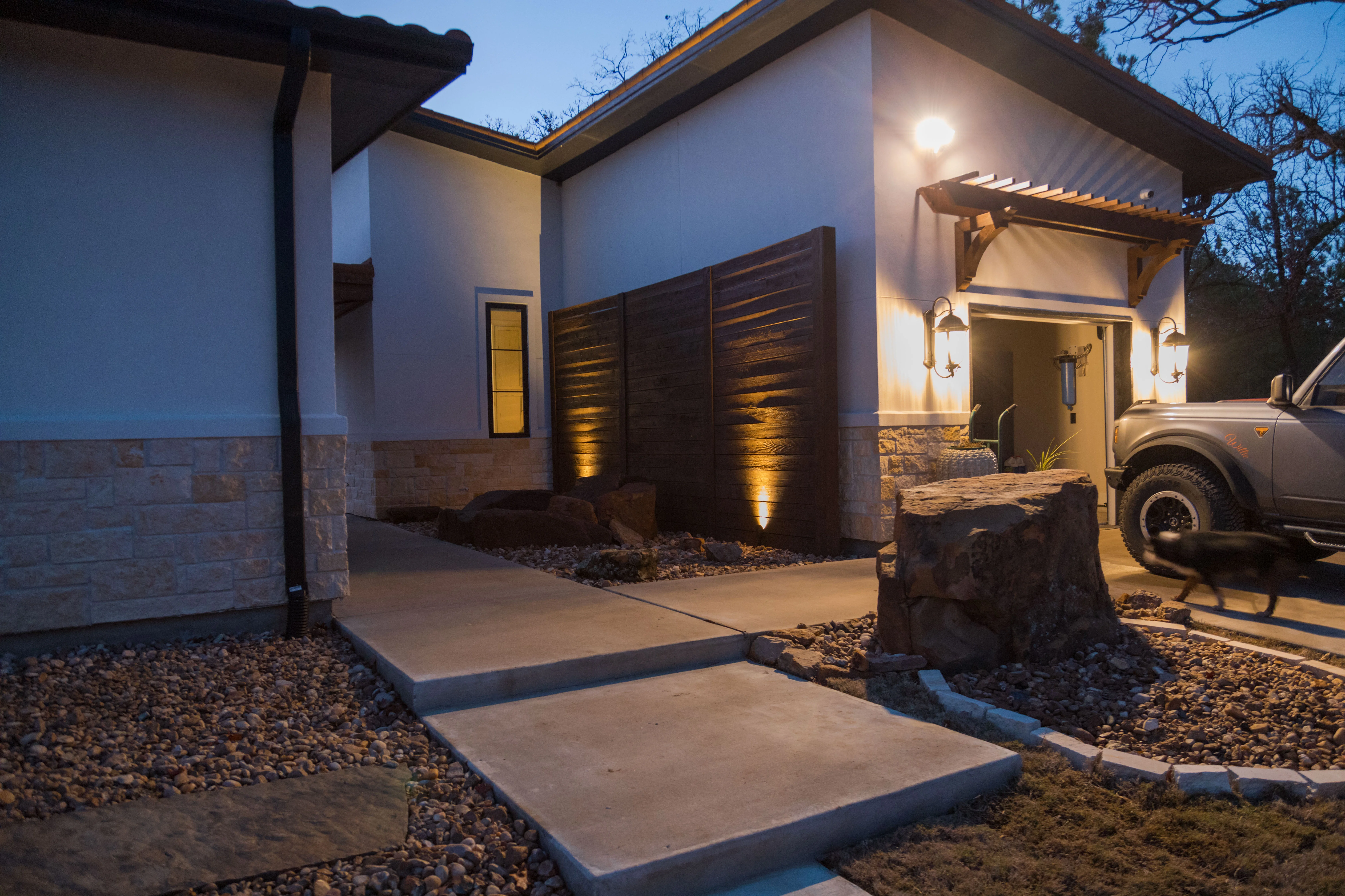 Illuminated modern home entrance with concrete walkways, river rock beds, and uplighting on dark wood screens at dusk — built by Landscaping Ninjas in College Station, TX