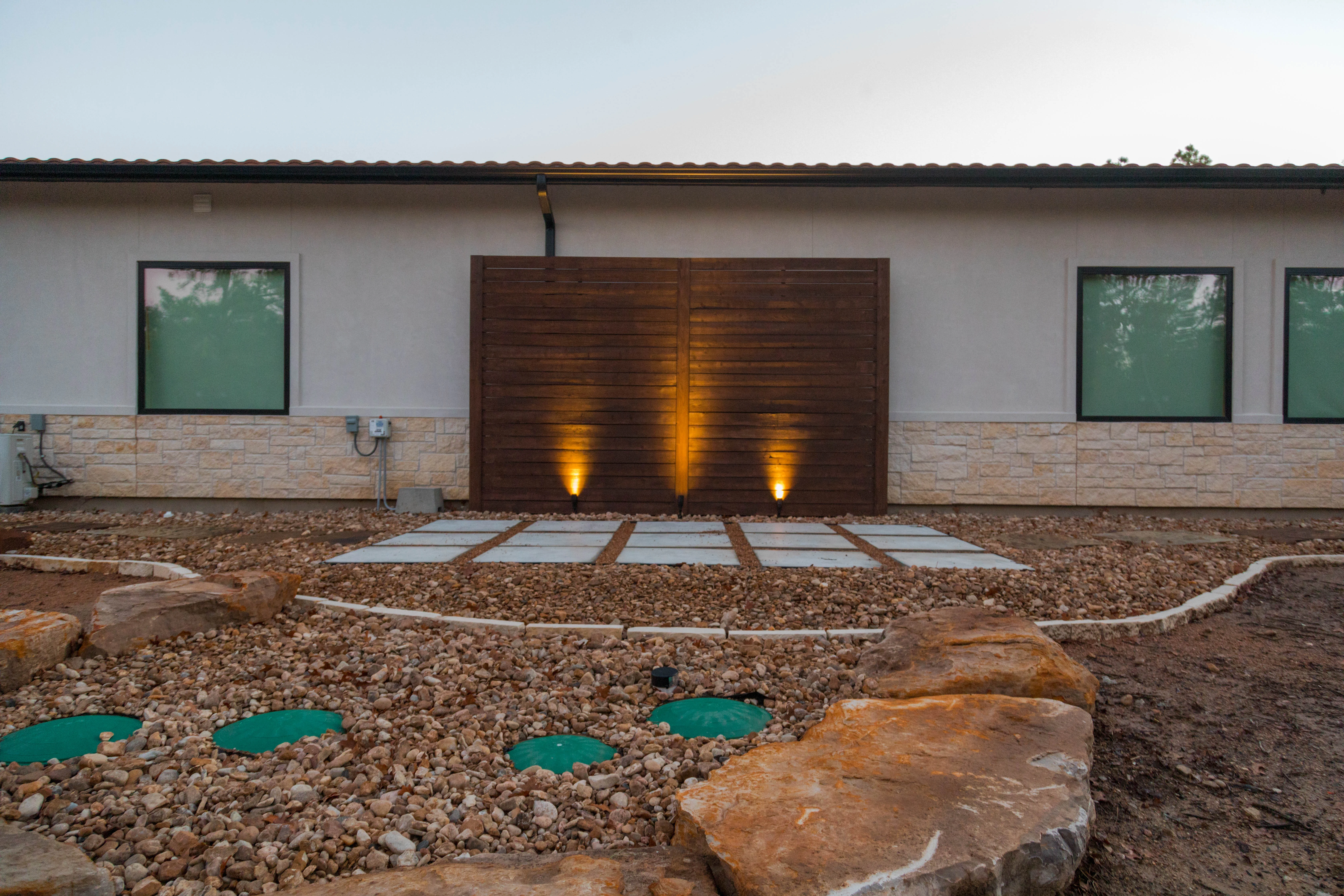 Modern home landscape with lit wood gate, gravel yard, and stone veneer foundation