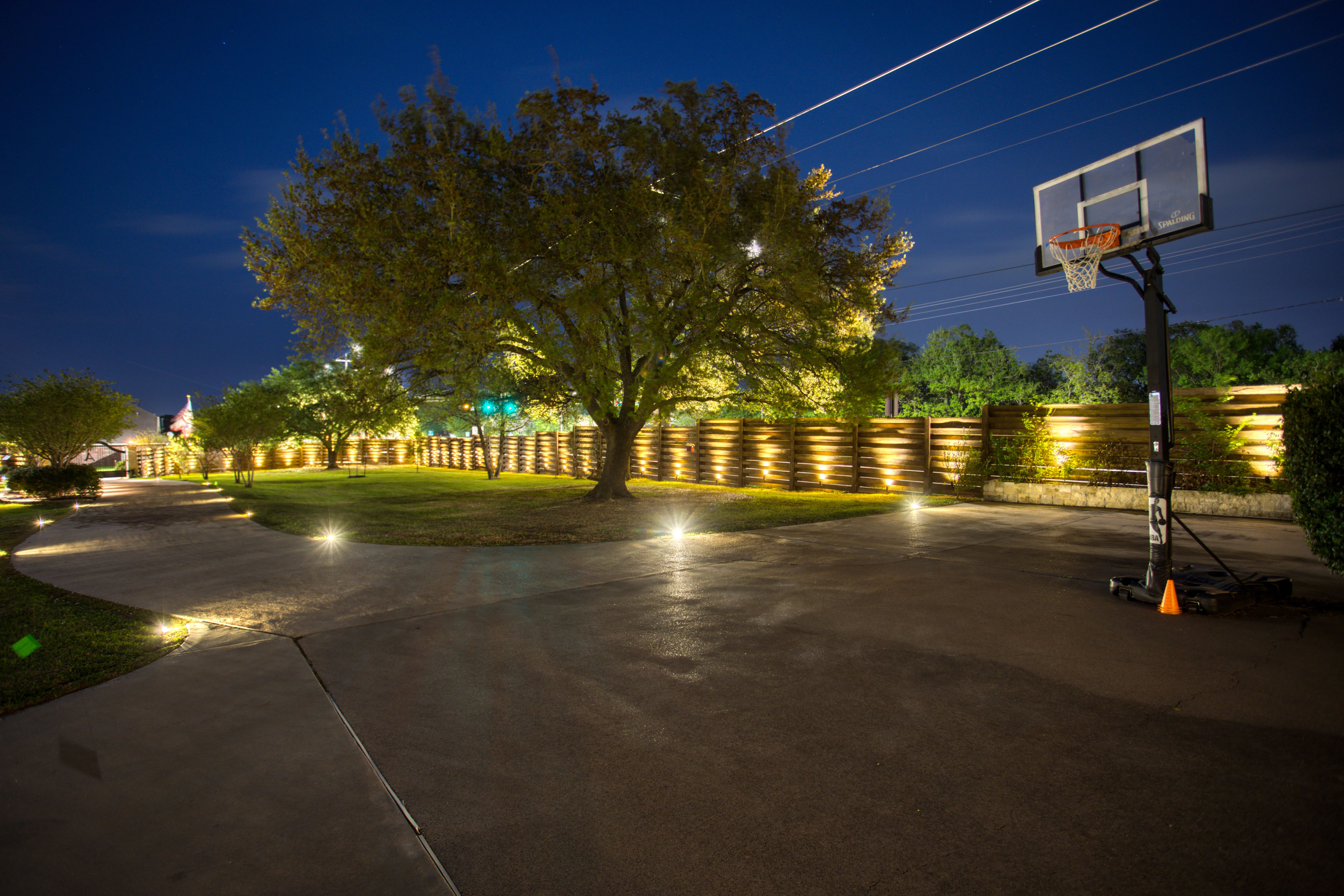 Illuminated residential landscape at night with lit fence, mature trees, and concrete patio