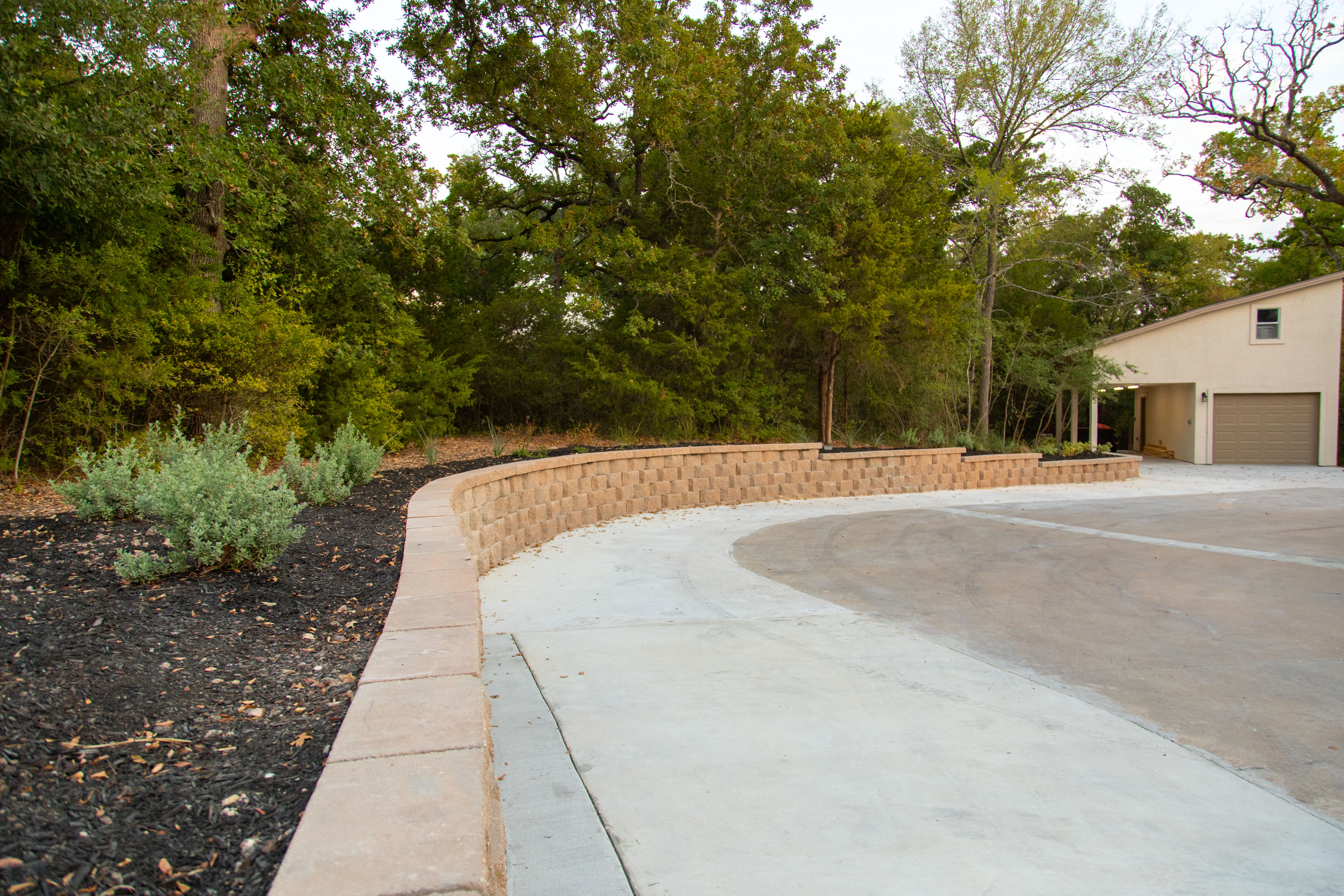 Curved brick retaining wall along concrete driveway with mature trees and landscaping
