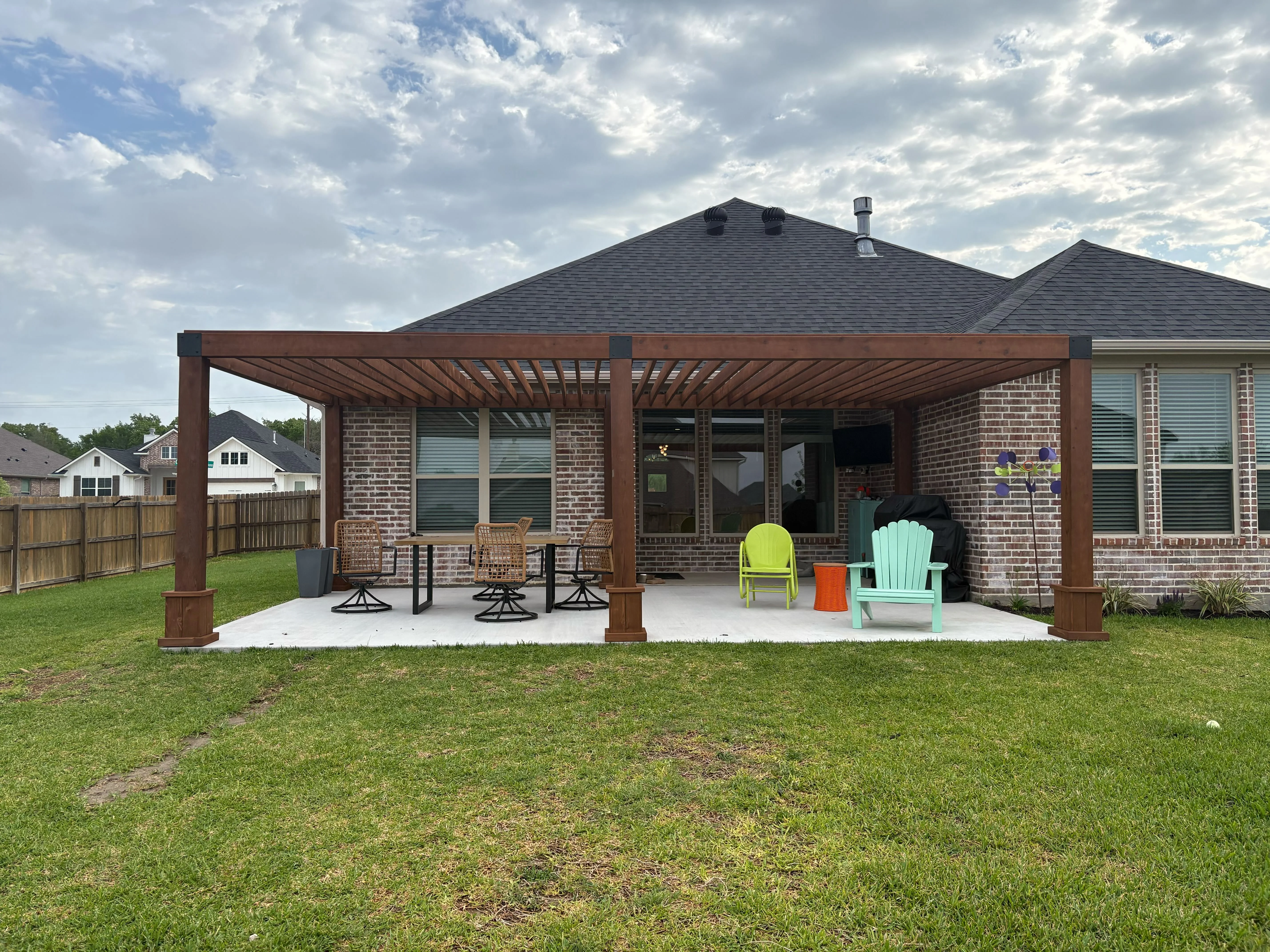 Brown pergola with louvered roof over composite patio beside brick home with green lawn