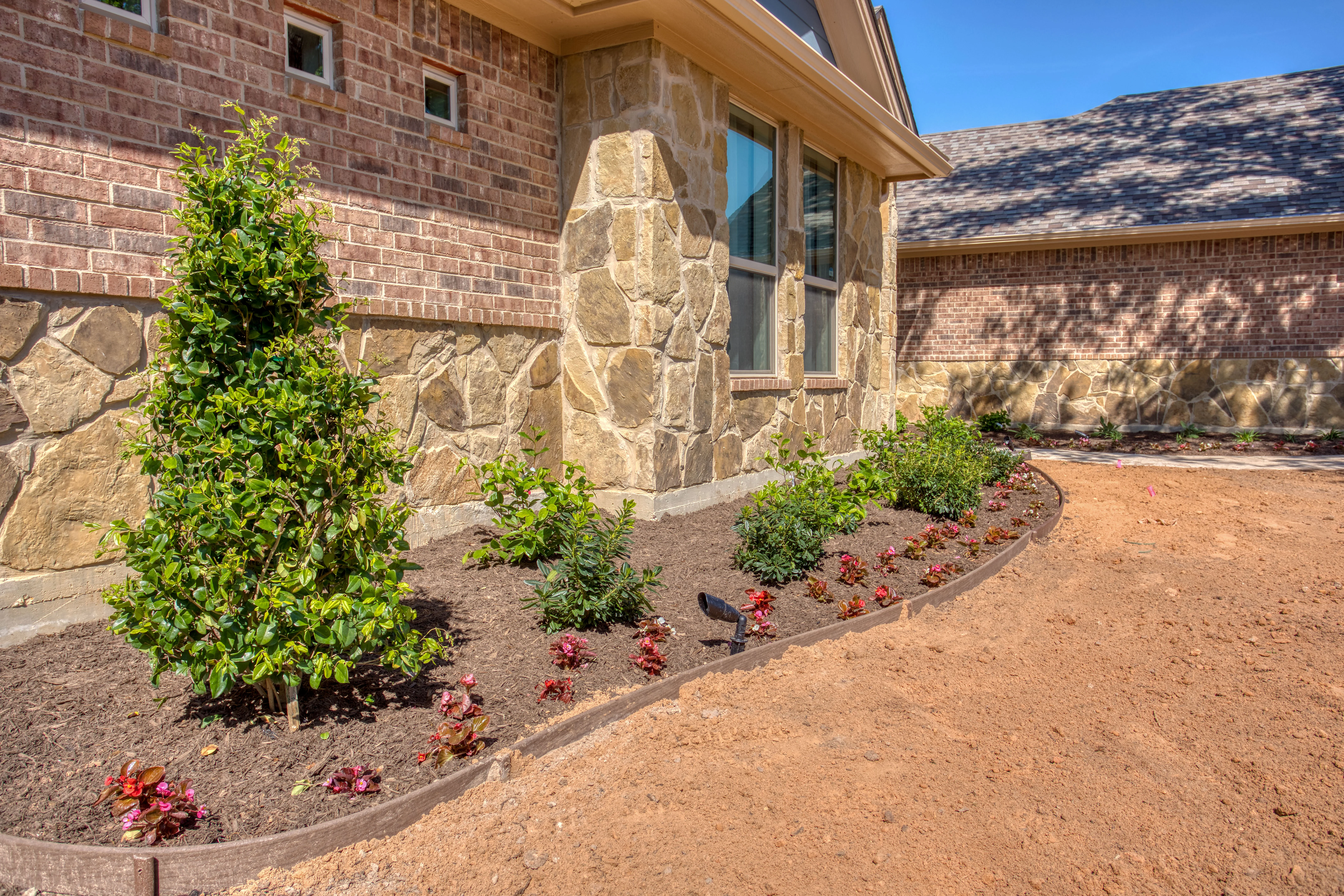 Newly installed landscape beds with mulch, shrubs, and flowering plants along brick and stone home foundation