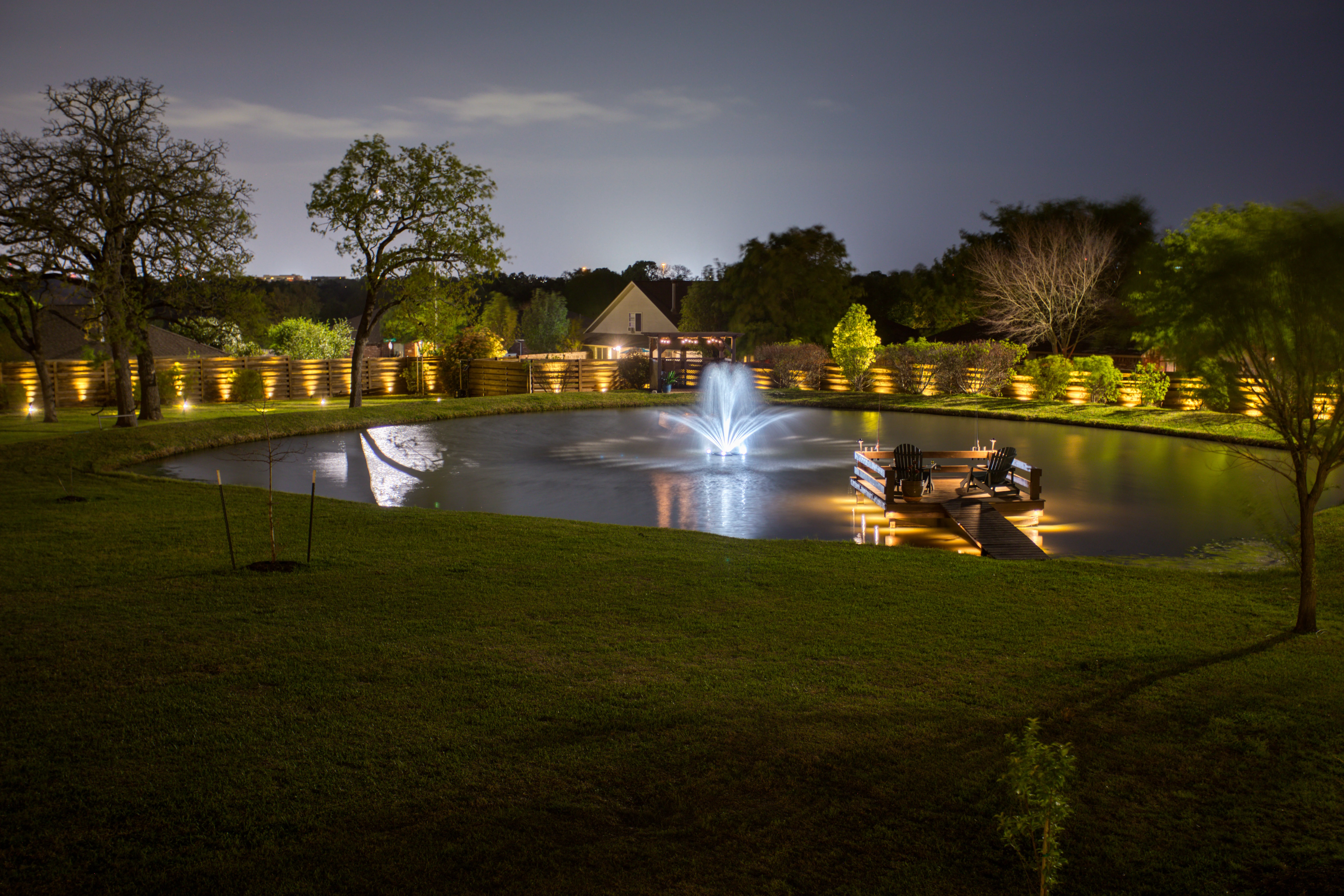 Nighttime view of illuminated residential pond with fountain, dock, and lit fence