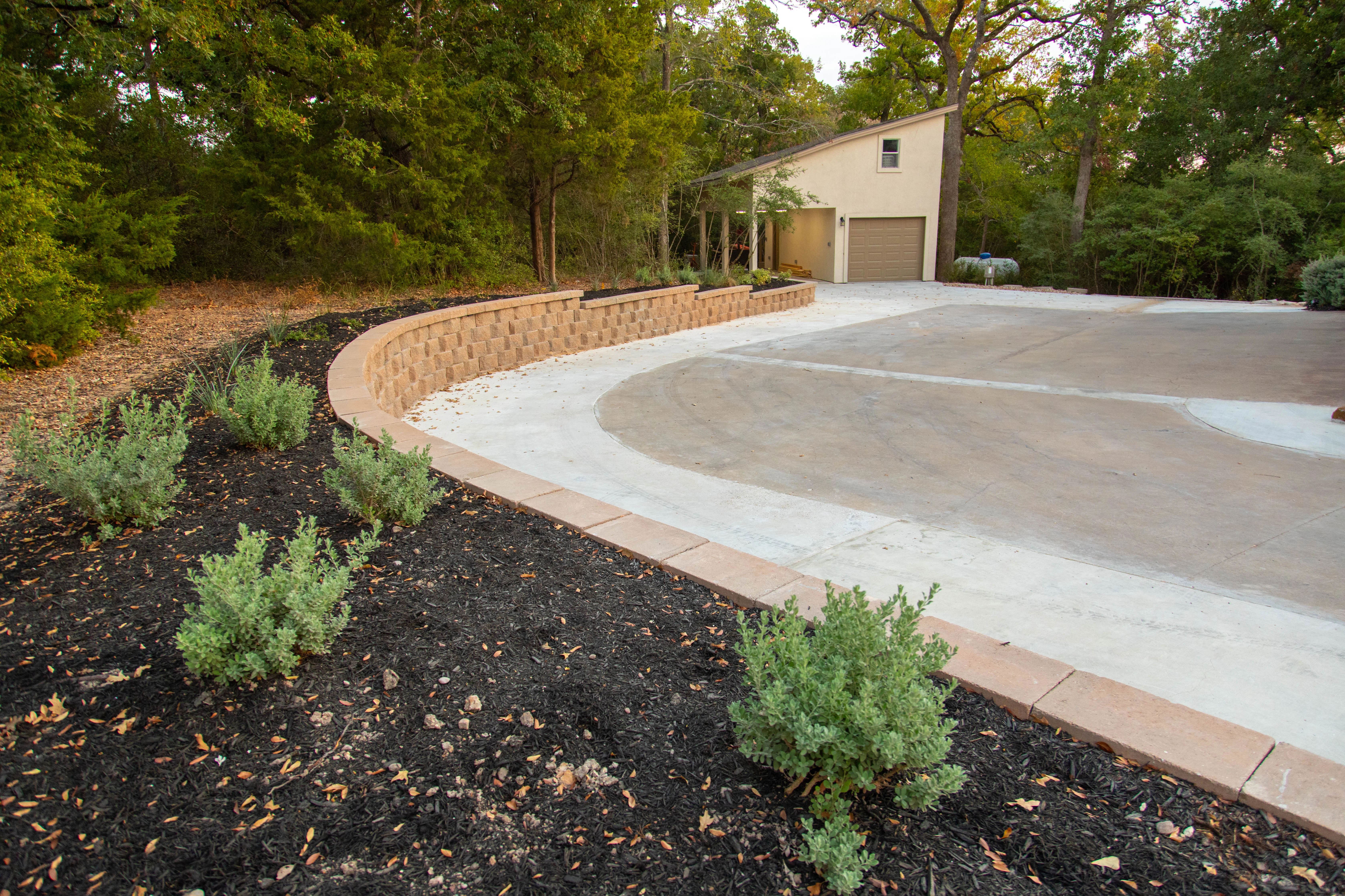 Curved tan retaining wall with concrete driveway and landscape plantings in wooded residential setting