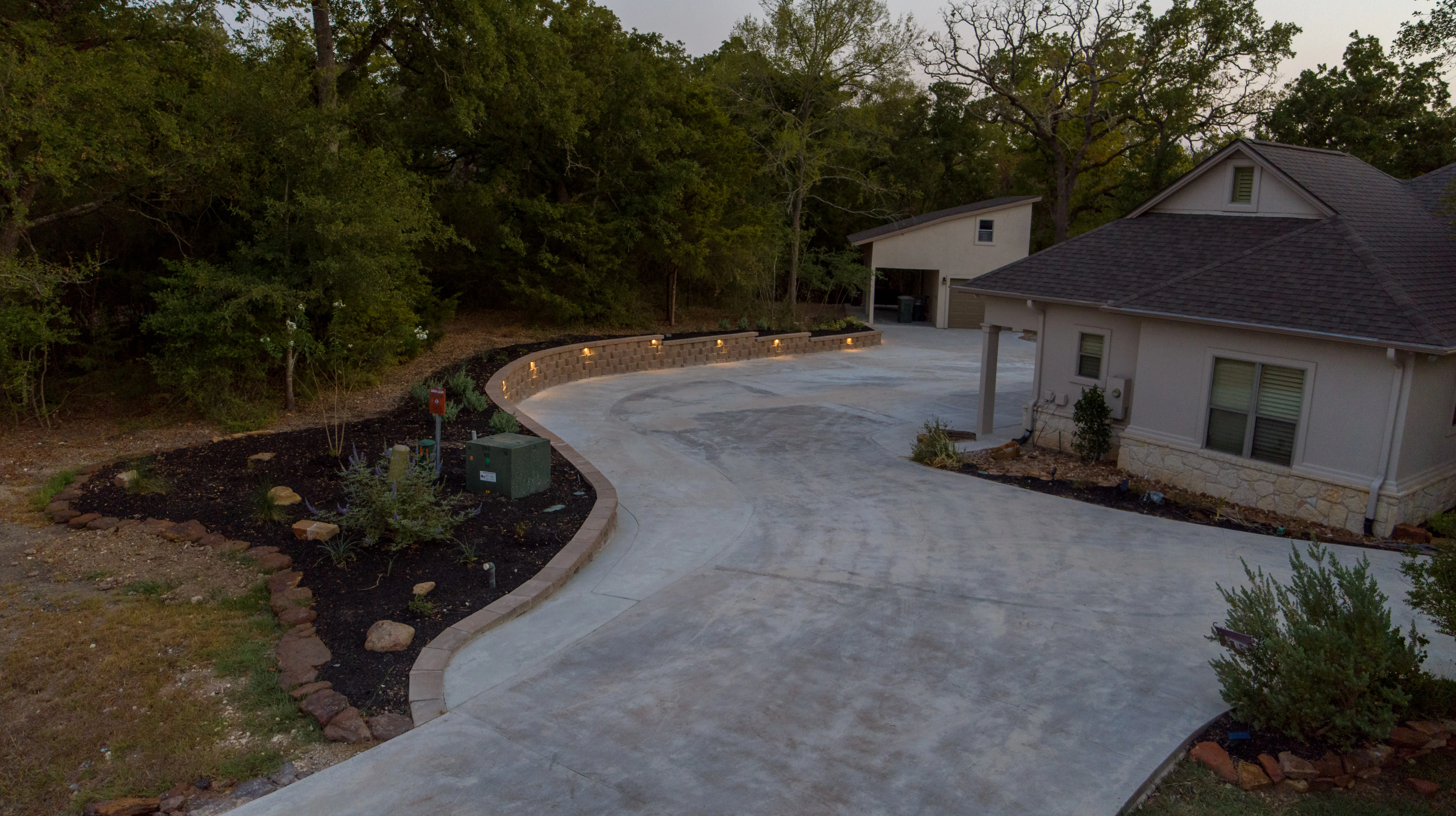 Curved concrete driveway with illuminated stone retaining wall and landscaped beds