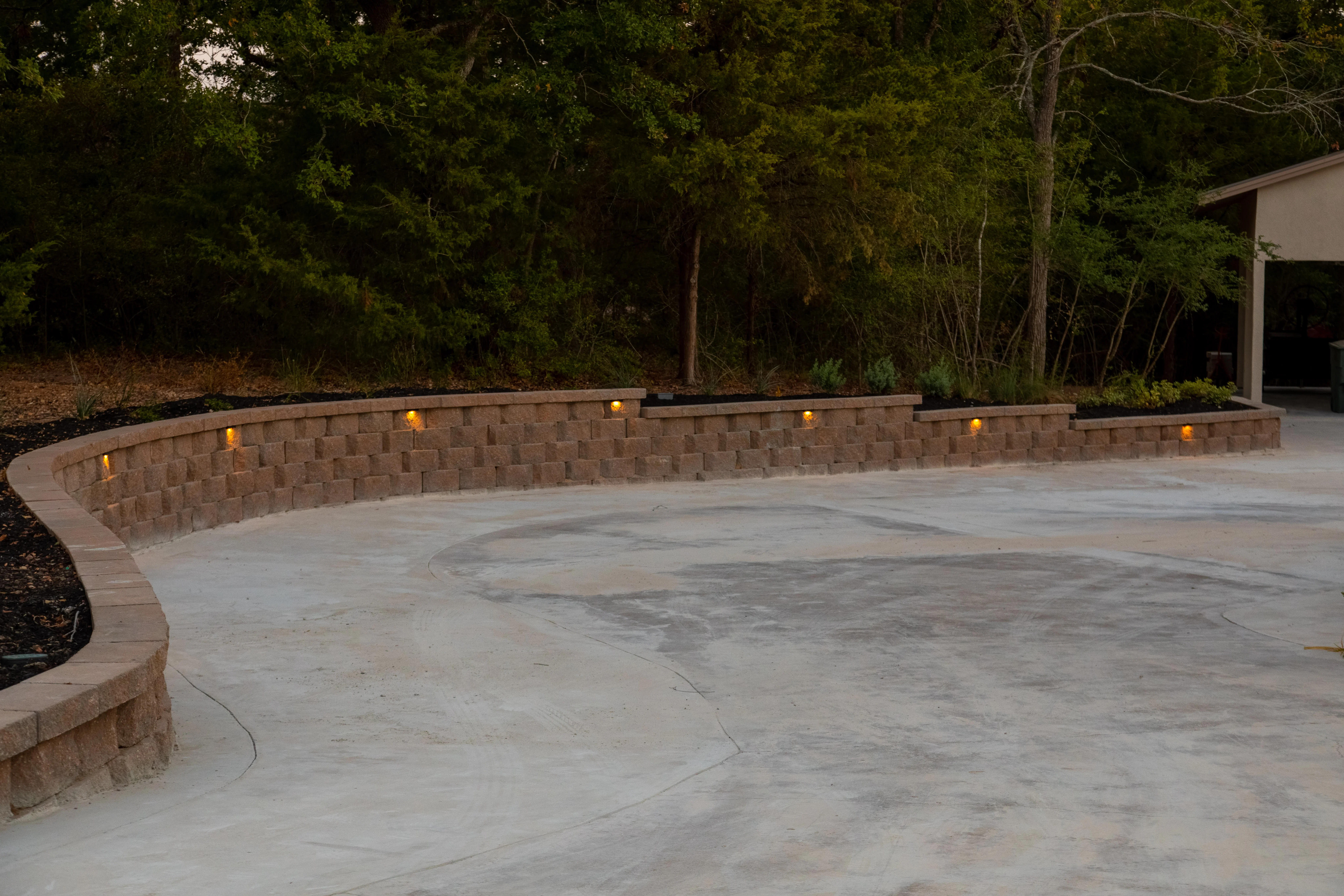 Curved tan retaining wall with uplighting overlooking concrete patio and trees